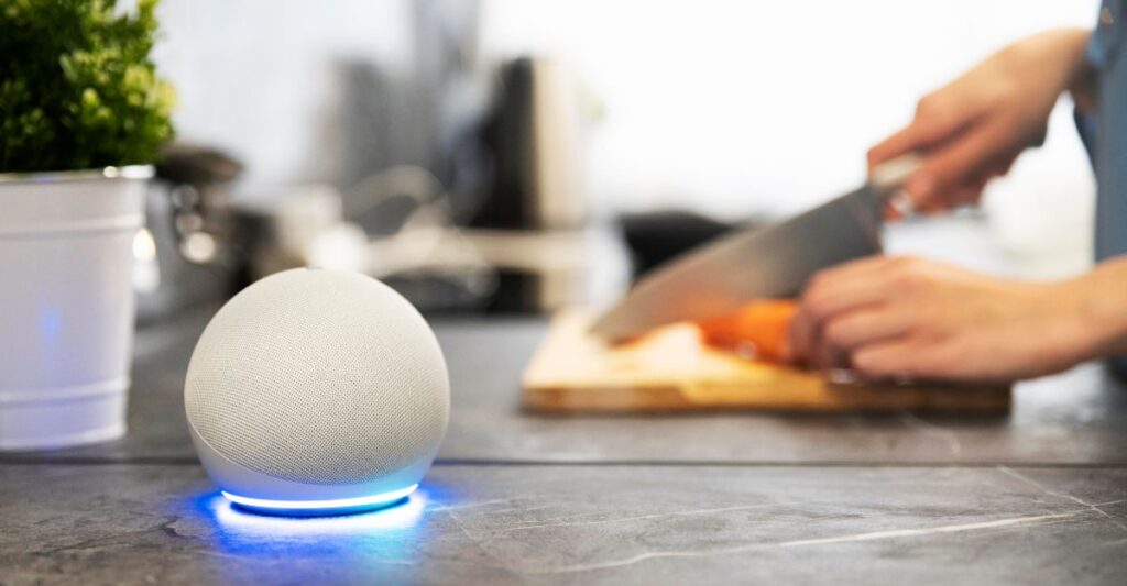 alexa cozinha dispositivo inteligente bancada.jpg Dispositivo inteligente com Alexa sobre bancada de cozinha, enquanto uma pessoa corta legumes ao fundo.