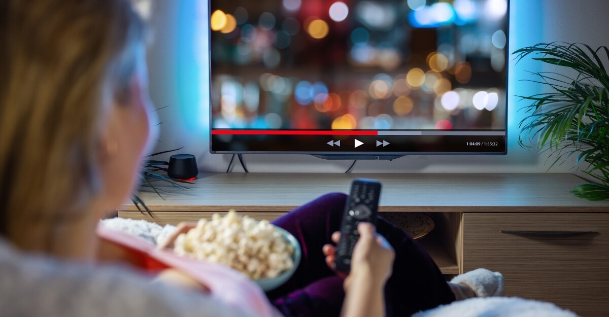 fire tv stick assistir filmes em casa 2025.jpg Mulher assistindo streaming com controle remoto e pipoca, usando o Amazon Fire TV Stick em uma TV inteligente moderna.